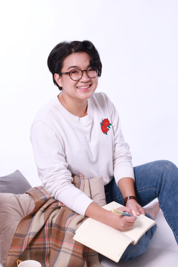 Didi, seated on the floor with pillows and a blanket, smiling, holding a pen to a blank notebook.